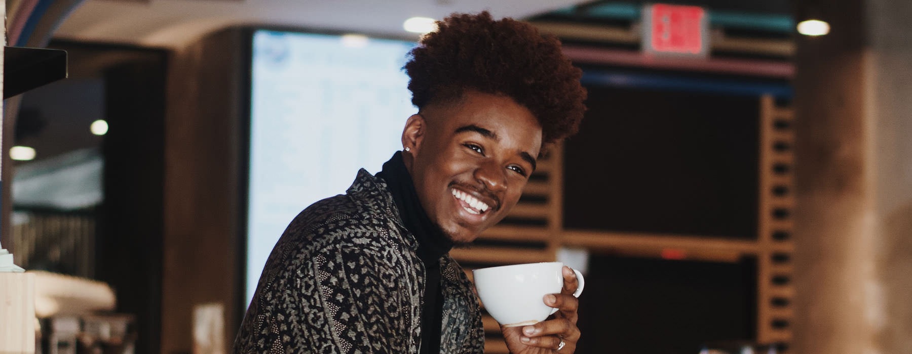 young man smiles at the camera as he holds a cup of coffee in local cafe